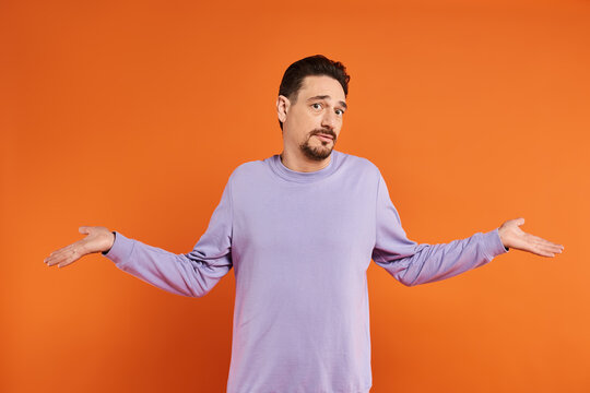 confused bearded man in purple sweater showing shrug gesture with his hands on orange background