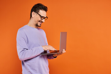 smiling man in eyeglasses and purple sweater using laptop on orange background, remote work