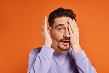 amazed man with beard in purple sweater hiding face with hand on orange background, playful mood