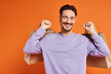 positive bearded man in casual attire holding shopping bags on orange background, consumerism