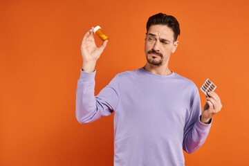 bearded man in purple sweatshirt holding bottle with pills and blister pack on orange background - obrazy, fototapety, plakaty