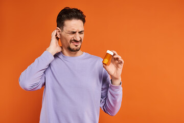 bearded man in purple sweatshirt holding bottle with pills on orange background, medication