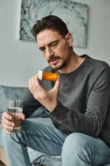 Fototapeta premium bearded man in grey jumper frowning and holding glass of water while looking at bottle with pills