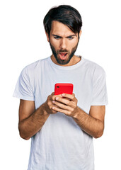 Hispanic man with blue eyes using smartphone typing message afraid and shocked with surprise and...