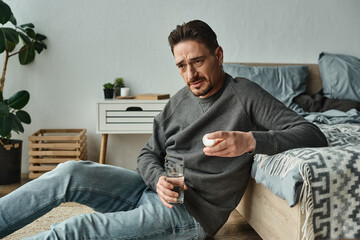 worried bearded man sitting on floor and holding glass of water and pills in bedroom, medication