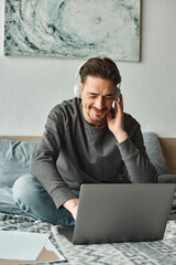 bearded man in wireless headphones holding documents and using laptop on bed, remote work