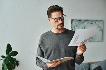 focused and bearded man in eyeglasses and grey sweater analyzing graphs while working from home