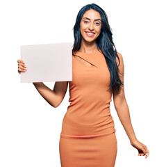 Beautiful hispanic woman holding blank empty banner looking positive and happy standing and smiling with a confident smile showing teeth