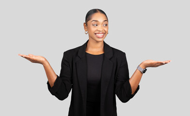 A smiling young woman in a black blazer presents a comparing gesture with both hands open