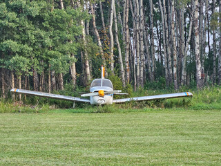 An old single propeller airplane sits abandoned in the green grass along the edge of a grass runway with a tree area behind it.