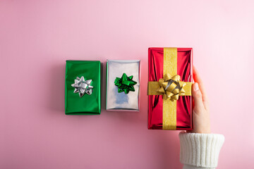 First person top view photo of female hands holding shiny red gift box over green surprise boxes with bow on isolated pastel pink background.