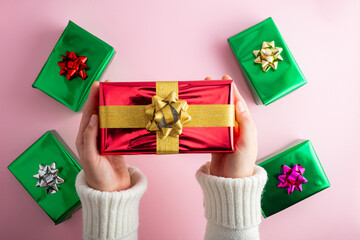 First person top view photo of female hands holding shiny red gift box over green surprise boxes with bow on isolated pastel pink background.