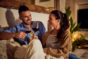 Two lovers sitting on the couch, covered in a blanket, holding glasses of wine, cheers.