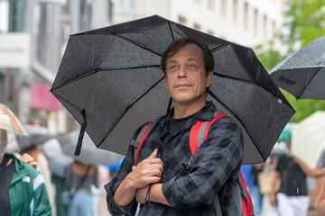 Street portrait of an elderly strong man under an umbrella in the rain, people pass by.