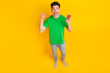 Full size photo of satisfied ecstatic guy wear stylish t-shirt denim pants hold smartphone win lottery isolated on yellow color background