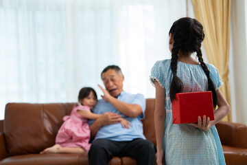 Little girl hiding present box behind back while making surprise elderly grandparents, Cheerful elderly man smiling and gesticulating while receives a surprise gift from granddaughter for celebration