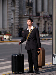 Portrait of handsome Chinese young man with black short hair wearing black blazer holding suitcase posing with modern city building background in sunny winter day, male fashion, cool Asian young man.