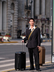 Portrait of handsome Chinese young man with black short hair wearing black blazer holding suitcase posing with modern city building background in sunny winter day, male fashion, cool Asian young man.
