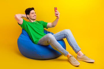Full length photo of cool man dressed green t-shirt jeans sit on pouf making selfie on smartphone...