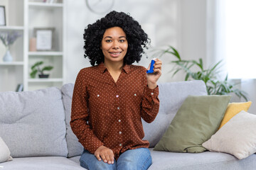 Portrait of a young smiling African American woman sitting at home on the couch in front of the camera, holding and showing asthma spray - obrazy, fototapety, plakaty