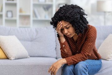 Upset young African American woman sitting on sofa at home, resting head on hand, feeling lonely, suffering from depression, thinking about solving problems