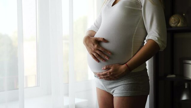 A pregnant woman strokes her belly with her hand. The girl's stomach itches. The child pushes in the stomach. Caring for the newborn. The wife is waiting for her husband near the window.