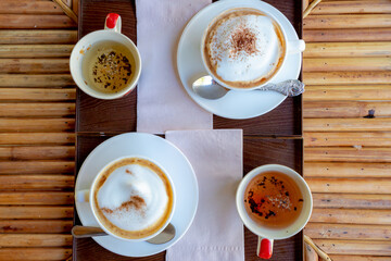 Barista concept, Selective focus of cups of cappuccino on bamboo table as background, Top view two cup of latte art coffee in white cup on wooden try or board served with cup of tea on the side.