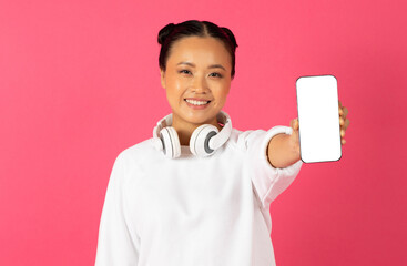 Cheerful young asian woman wearing headphones demonstrating smartphone with blank screen