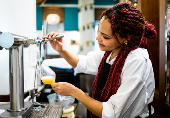 A dark-skinned woman in uniform working inside a coffee shop. The African girl with braided hair is...