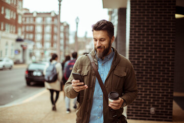 Man walking with coffee and using smartphone in city street - Powered by Adobe