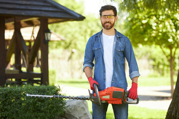 Confident gardener in protective glasses smiling, while posing with electric hedge trimmer...