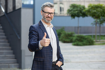 Smiling mature businessman in stylish suit showing thumbs up gesture outside of a contemporary...