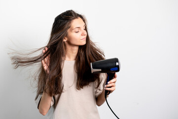 A content young woman is seen styling her flowing brown hair with a blow dryer against a neutral...