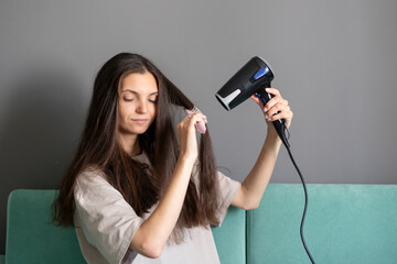 A woman comfortably styles her long dark hair using a hairdryer and round brush in a home setting, showcasing a common beauty routine.