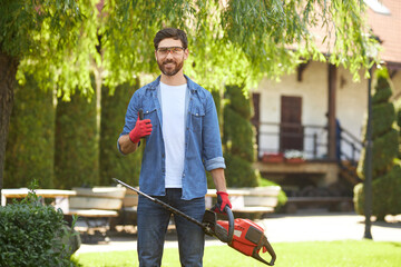 Handsome gardener in gloves showing thumb up, while standing with electric hedge trimmer in garden....