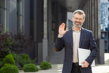 Cheerful senior businessman with a laptop greeting with a wave outside an urban office, symbolizing approachability and professionalism.