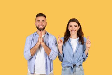 Hopeful couple with praying gesture and crossed fingers, optimistic on yellow