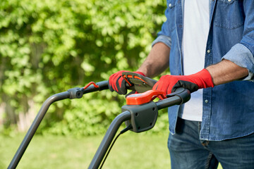 Fototapeta na wymiar Crop view of gardener in casual outfit mowing lawn with modern lawn mower in summer. Close up view of muscular male hands in gloves on lawn mower handle against hedge background. Concept of gardening.