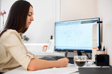 Portrait of a remote worker. A beautiful woman sits at a table in front of a computer and designs a building. Confident young lady freelancer working from home.