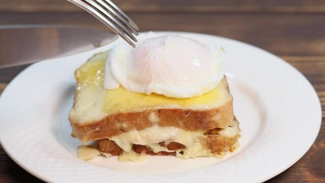 Close up view of Poached egg spreads over the sandwich in slow motion. Croque madame, grilled sandwiches with sliced ham, melted cheese, dijon mustard and poached egg in bechamel sauce.