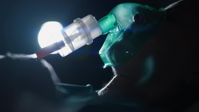 A female patient in medical mask is breathing oxygen support in a hospital next to heart rate machine. Unhealthy woman in coma. Getting mechanical ventilation. Concept of pandemic and therapy.