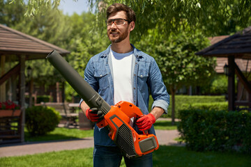 Handsome male worker in casual apparel using modern gasoline cordless leaf blower on backyard....