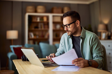 A focused adult male accountant working from home on his laptop and holding documents in his hand.