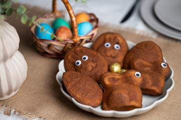 Close-up of bowl full of funny muffins shaped as bunnies and eggs with eyes. Multi colored Easter eggs in wicker basket and vase with green plant on served table