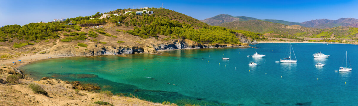 Panoramic view of Playa del Borr&oacute; in Colera, with crystal clear waters and anchored yachts, surrounded by green hills and coastal homes on a sunny day.