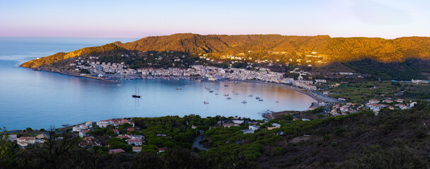 Panoramic view of Port de la Selva Bay at twilight, with boats anchored in calm waters, illuminated...