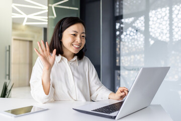 Professional young Asian businesswoman in a virtual meeting, waving with a friendly smile at her laptop in a bright office setting.