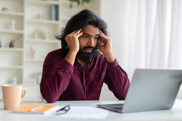 Concerned young indian man looking at laptop screen and holding his temples