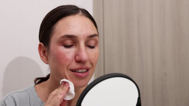 millennial young woman is doing at home facial procedure, remove makeup with hydrophilic oil and cotton disk,massaging face with roller and cleaning with ultrasound device.girl using mirror.