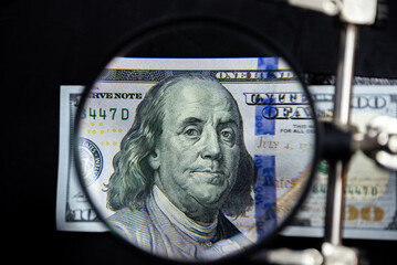 100 US dollar note with Benjamin Franklin face portrait magnified with a magnifying glass on a dark background. Monetary wallpaper.